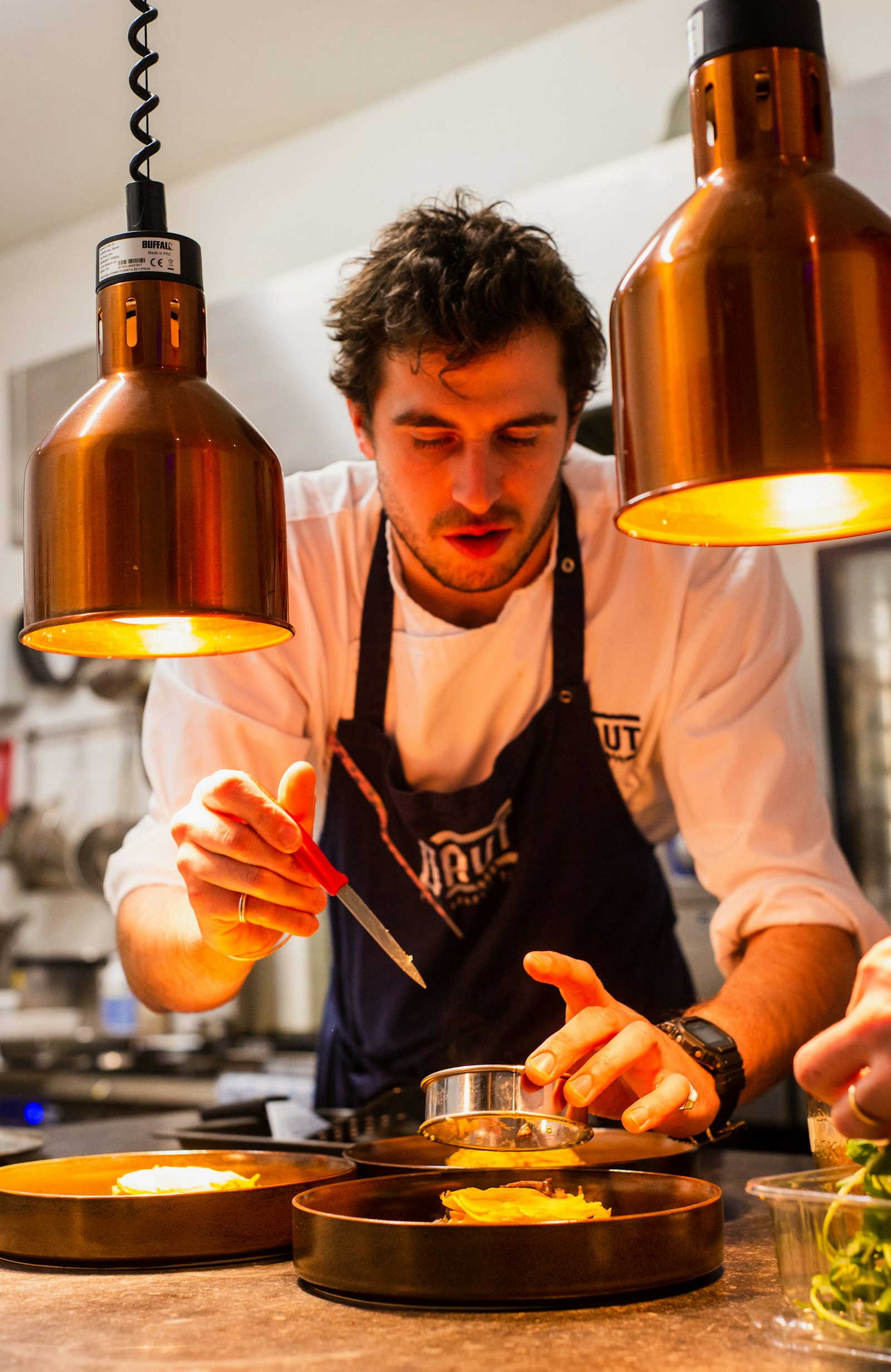 A chef finishing a plate under copper heat lamps